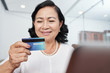 © DragonImages - Asian senior woman looking at her credit card in her hand while sitting at the table in front of laptop