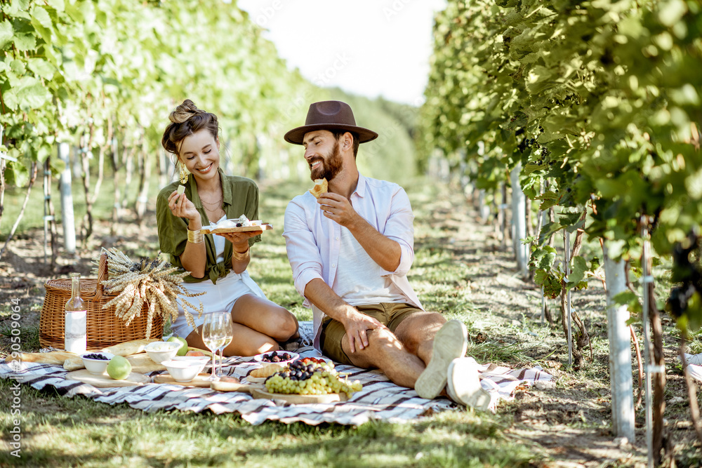 Beautiful couple having romantic breakfast with lots of tasty food and wine, sitting together on the picnic blanket at the vineyard on a sunny morning - obrazy, fototapety, plakaty 