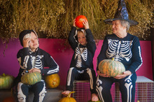 Elderly Witch Grandmother Is Sitting In Barn With Her Grandchildren. Children And Old Woman Are Dressed In Skeleton Costumes. Halloween Eve With Pumpkins In Hands