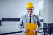 © Dusan Petkovic - A businessman in a grey suit with a safety gear on his head standing and looking directly into the camera with a yellow file in his hands