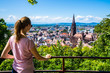 Germany, Beautiful young tourist girl standing above skyline of freiburg im breisgau city and famous muenster cathedral in summer surrounded by green trees