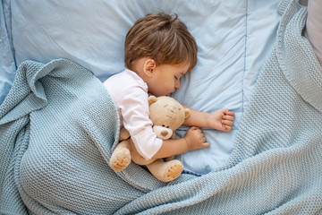 Wall Mural - Photo of baby boy sleeping together with teddy bear. His favorite napping spot. Adorable kid boy after sleeping in bed with toy. Boy sleeping on bed with teddy bear. Sleepyhead