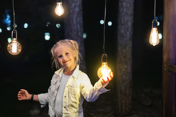 Cute adorable caucasian blond girl portrait smiling and holding in hand one of hanged edison light bulb at forest outdoor. Right solution choice concept