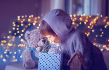 Christmas Mood. Cute Little Excited Child With Teddy Looking Inside Gift Box With Christmas Toys And Light From It With Garland Lights Bokeh At Background At Home.  Purple Violet Magic Toning.
