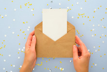 Female's Hands Holding Kraft Paper Envelope Letter With Blank White Card Mockup Over Pastel Blue Background With Golden Confetti Stars. Christmas, New Year, Winter Holidays And Birthday Concept.