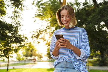 Wall Mural - Image of smiling woman typing on cellphone while resting in park