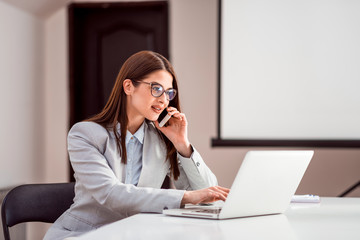 Wall Mural - Cute young female adult with glasses talking on phone while working on laptop.