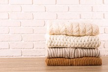 Bunch Of Knitted Pastel Color Sweaters With Different Knitting Patterns Perfectly Folded In Stack On Brown Wooden Table, White Brick Wall Background. Fall Winter Season Knitwear. Close Up, Copy Space.