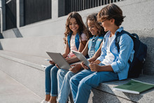 Happy Group Of Multiethnic School Kids Sitting And Using Digital Devices Outdoors