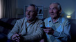 © motortion - Happy aged men watching tv at night holding remote control, nursing home leisure