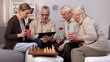 © motortion - Elderly man showing friends new project on tablet pc, chess play brake, fun
