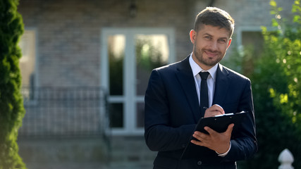 Broker filling in papers smiling to camera, preparing document for selling house