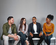 Multiracial happy young friends sitting relaxed and cheerful on chair in queue against gray wall - real authentic people being included and feeling a sense of belonging