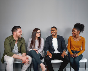 Poster - Multiracial happy young friends sitting relaxed and cheerful on chair in queue against gray wall - real authentic people being included and feeling a sense of belonging