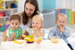 © Oksana Kuzmina - Babies having lunch in kindergarten. Kids eat healthy food in creche. Carer with little children sitting at dinner table in canteen