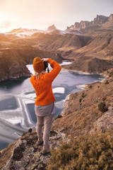 Wall Mural - Portrait from the back of the girl traveler photographer in an orange sweater and hat with a camera in hand in the mountains against the background of a frozen mountain lake. Photo travel concept