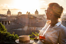 Young Woman Tourist Fashion White Dress With Spritz Cocktail In Front Of Panoramic View Of Rome Cityscape From Campidoglio Terrace At Sunset. Landmarks, Domes Of Rome, Italy.