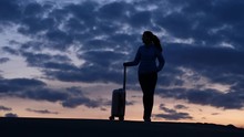 Tourist Woman Stay Alone At Top, Hold Handle Of Small Trolley Case. Shaded Figure Against Dark Clouds On Evening Sky. Twilight Hour, Traveller Wait At Road Side.