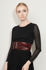 Cropped front view shot of a blond girl, wearing black dress with transparent sleeves and red snake print wide belt, decorating with two buckles. Girl is posing on a white background. 