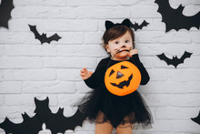 A Little Girl In Black Cat Costume Bites Pumpkin Basket On Bats Background, Trick Or Treat, Halloween Concept.