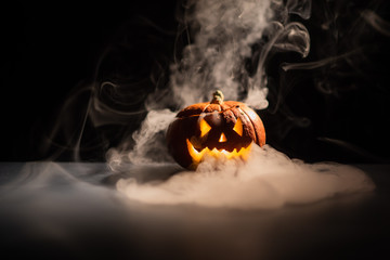 Halloween, orange pumpkin with a scary luminous face on a dark background. Gray thick smoke comes out. Close-up of jack-o'-lantern on the eve of all saints.