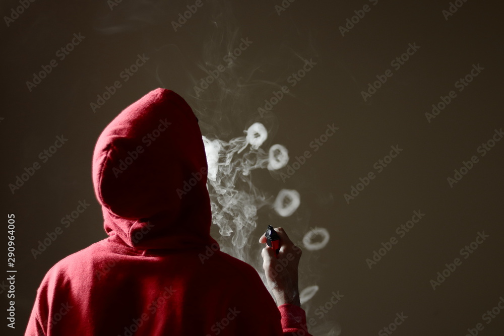 Young male in red hoodie vaping smoking, blows multiple smoke rings while  holding a vape in hand, isolated rear view Stock Photo