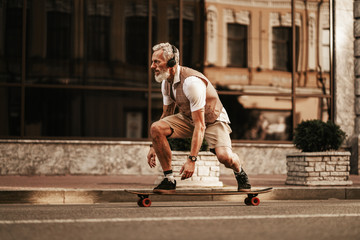 Portrait of bearded hipster man in white shirt on city street. Stylish happy model ride on longboard near road on buildings background