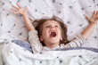 © sementsova321 - Close up portrait of cheerful little girl lying in bed under blanket with dandelion, charming kid wearing pajama, spreading her arms, keeps mouth openes, screams something happily. Childhood concept.