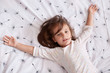 © sementsova321 - Top view of little girl laying in bed and spreading her arms on white cover with dandelion, charming kid with dark hair wearing pajama, looking at camera, relaxing after hard day. Children concept.