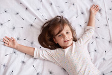 Top View Of Little Girl Laying In Bed And Spreading Her Arms On White Cover With Dandelion, Charming Kid With Dark Hair Wearing Pajama, Looking At Camera, Relaxing After Hard Day. Children Concept.