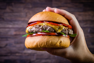 Hand holding a craft beef burger with cheese, italian peperoni, tomato and basil leafs on rustic background.