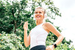 © Adamov - Portrait of cheerful young woman smiling and listening to music while jogging in the park. Motivated sportswoman in headphones running outdoors. Horizontal shot. Low angle. Greenery in the background