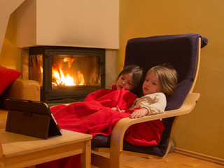 two little sisters sit by the fireplace on a winter evening and watch cartoons on a tablet