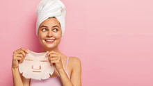 Positive Young Female Model Holds Beauty Mask, Applies On Face For Rejuvenating, Looks Aside, Wears White Towel On Head, Keeps Gaze Right, Poses Over Pink Background, Copy Space For Your Promotion