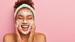 © wayhome.studio  - Close up shot of cheerful African American woman massages cheeks with both hands, cleans face with foam, has eyes shut, shows white teeth, naked body, wears headband, isolated on pink background