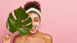 Studio shot of delighted woman with curly hair, applies cleansing foam on face, holds green plant leaf, peels complexion with foam, smiles broadly, poses over pink background, empty space for advert