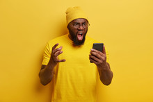 Annoyed Black Man With Thick Beard Shouts Angrily, Has Furious Face Expression, Gets Unpleasant News, Wears Vivid Yellow Hat And T Shirt, Poses Indoor, Being Angry With Customer Service, Stands Indoor