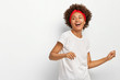 Photo of pretty dark skinned woman stands sideways, feels energized dances actively to music wears red headband casual white t shirt poses indoor. Horizontal shot of lovely African American girl moves