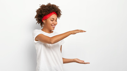 Poster - Horizontal shot of happy dark skinned curly haired woman shows height of something, gestures with both hands, demonstrates size of package, dressed in casual wear, isolated over white background