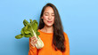 Indoor shot of delighted young woman returns from grocers shop, holds fresh green bok choy, keeps to healthy diet, wears orange jumper, isolated over blue background. Vegetables, vegetarian, calories