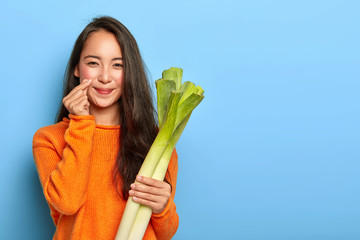 Photo of lovely Asian woman makes korean like hand sign, holds green leek bought in grocers shop, wears orange sweater, going to cook vegetarian dish, poses over blue wall with blank copy space