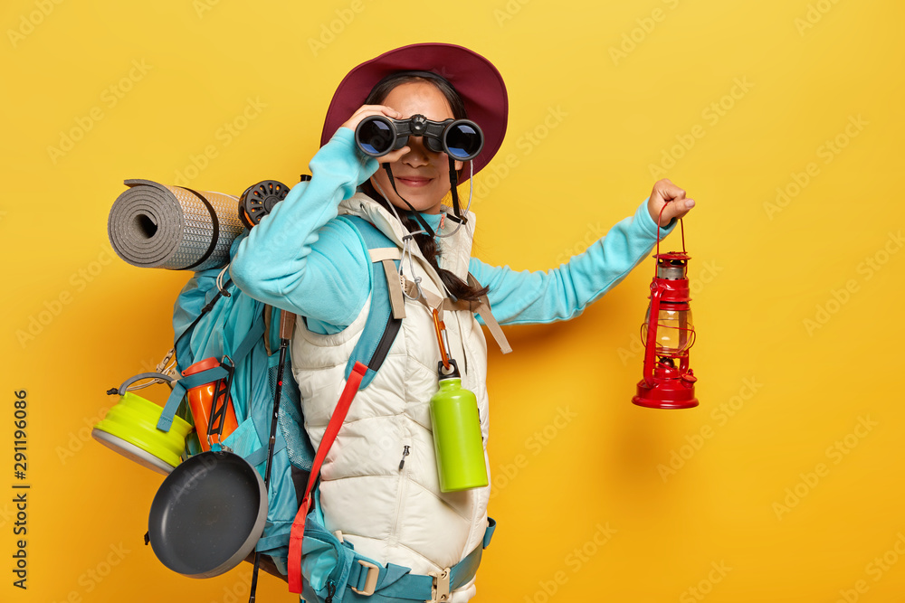 Horizontal shot of curious female backpacker explores tourist destination, uses binoculars, dressed in active wear, holds kerosene lamp carries travelling items with rucksack isolated over yellow wall - obrazy, fototapety, plakaty 