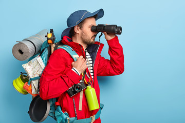 Photo of male explorer dressed in casual wear, keeps binoculars near eyes, wears hat and jacket, hikes in mountains, isolated over blue background copy space area for your advertisement. Tourist hiker