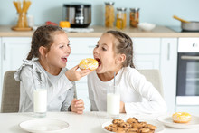 Portrait Of Cute Twin Girls Eating Donuts With Milk In Kitchen