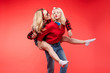© Вячеслав Косько - Stock studio portrait of two cheerful and positive trendy girlfriends or sisters having fun. Girl in sunglasses making bunny ears to her friend with her hand while sitting on her back. Isolate on red.