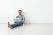 © satura_ - Positive young man in casual clothes surfing the Internet in search of new housing sitting on the floor in an empty room. The concept of finding an apartment using the Internet and a laptop. Copyspace