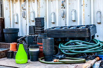 Accumulation of objects on a work table with a rusty metal wall with neon lamps in a greenhouse with hoses, spray bottle, flower pots, plastic containers for plants and flowers for recycling