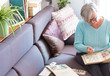 © luciano - An elderly person looks with interest at his old stamp collection. Senior female with gray hair. Sitting on the couch with cell phone and tablet. White wall and books in the background
