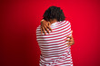 © Krakenimages.com - Young afro man with dreadlocks wearing striped t-shirt standing over isolated red background Hugging oneself happy and positive from backwards. Self love and self care