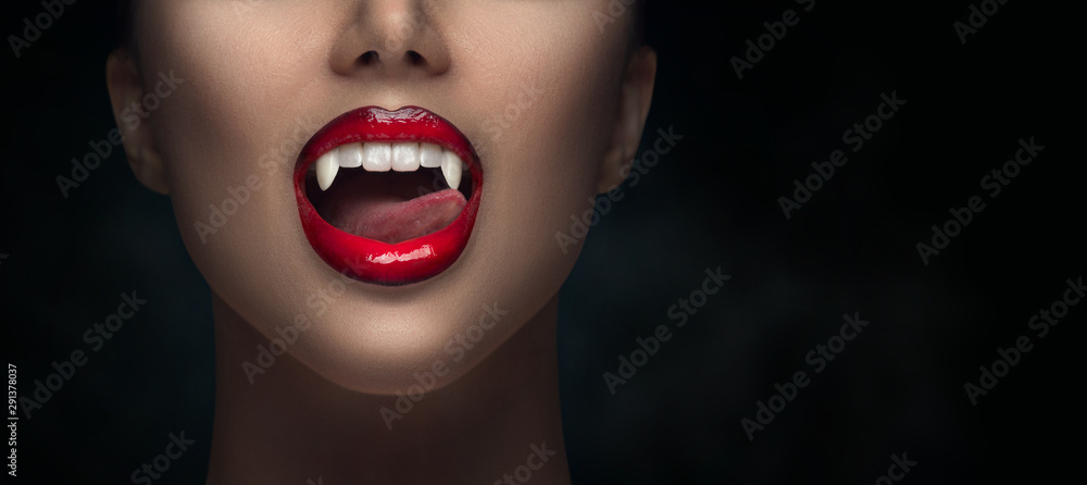 Sexy Vampire Woman's red bloody lips close-up. Vampire girl licking fangs with tongue. Fashion Glamour Halloween art design. Close up of female vampire mouth, teeth. Isolated on black background - obrazy, fototapety, plakaty 